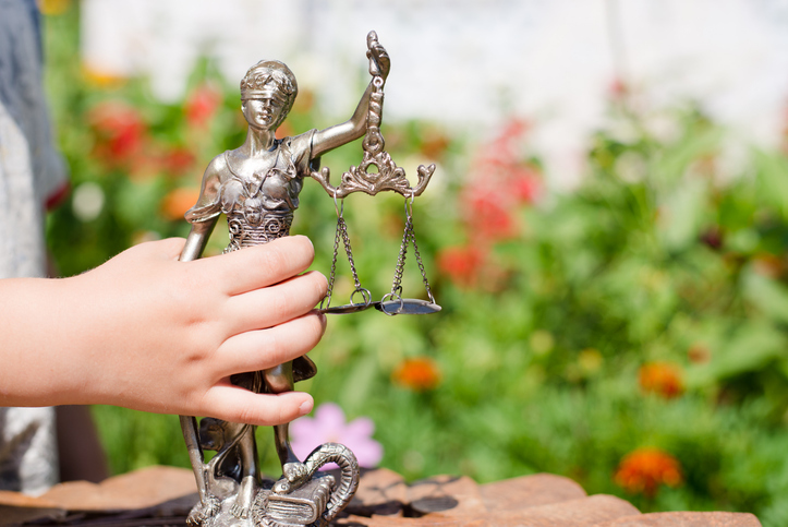 A child’s hand holds a metallic Lady Justice figurine outdoors, showing the statue’s scales and detailed armor, with a soft, colorful garden background.