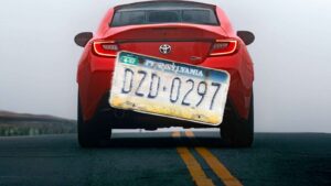 Back of a red car driving down the road with an enlarged license plate over the back.